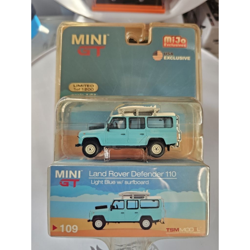 Mini GT #109 MiJo Exclusives Land Rover Defender 110 in light blue with a white top and two surfboar